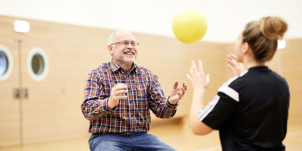 Eine Therapeutin wirft einem sitzenden älteren Patienten einen gelben Ball zu. Dieser fängt ihn gut gelaunt.