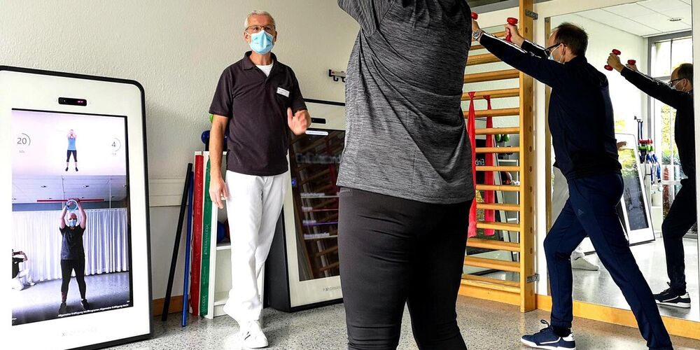 Christoph Schneider steht nehmen dem Pixformance Bildschirm und gibt zwei Patienten Anweisungen, die die angezeigten Übungen kopieren.