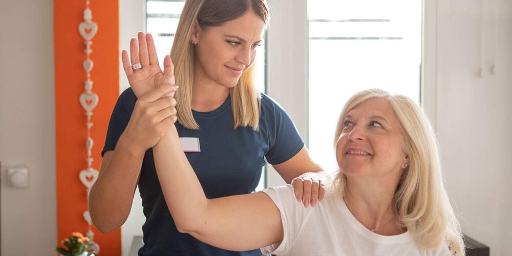 Attraktive Physiotherapeutin steht seitlich hinter der Patientin und behandeln deren Schulter. Beide sehen sich an.