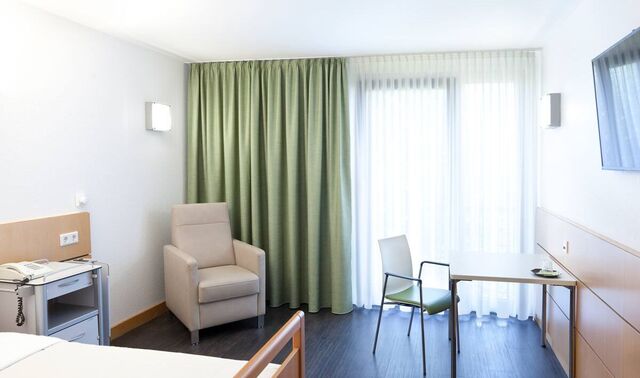Blick in das Einzelzimmer. Dunkler Holzboden, beiger Sessel, heller Tisch und Bett, hellgrüner Vorhang, viel Licht