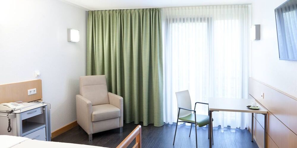 Blick in das Einzelzimmer. Dunkler Holzboden, beiger Sessel, heller Tisch und Bett, hellgrüner Vorhang, viel Licht