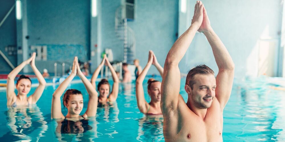 Ein Wassergymnastik-Kurs: Der muskulöse Kursleiter ist im Vordergrund zu sehen. Er streckt seine Arme in die Höhe. Dahinter machen vier Kursteilnehmerinnen die Übung nach.