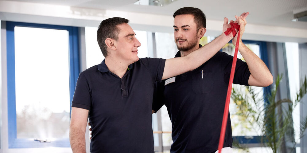 Junger Physiotherapeut unterstützt den Patienten bei einer Armübung mit Theraband.